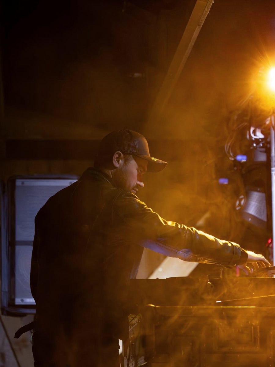 DJ Daniel Wulz mit Kappe am Pult, im Gegenlicht und Nebel in warmem Orange