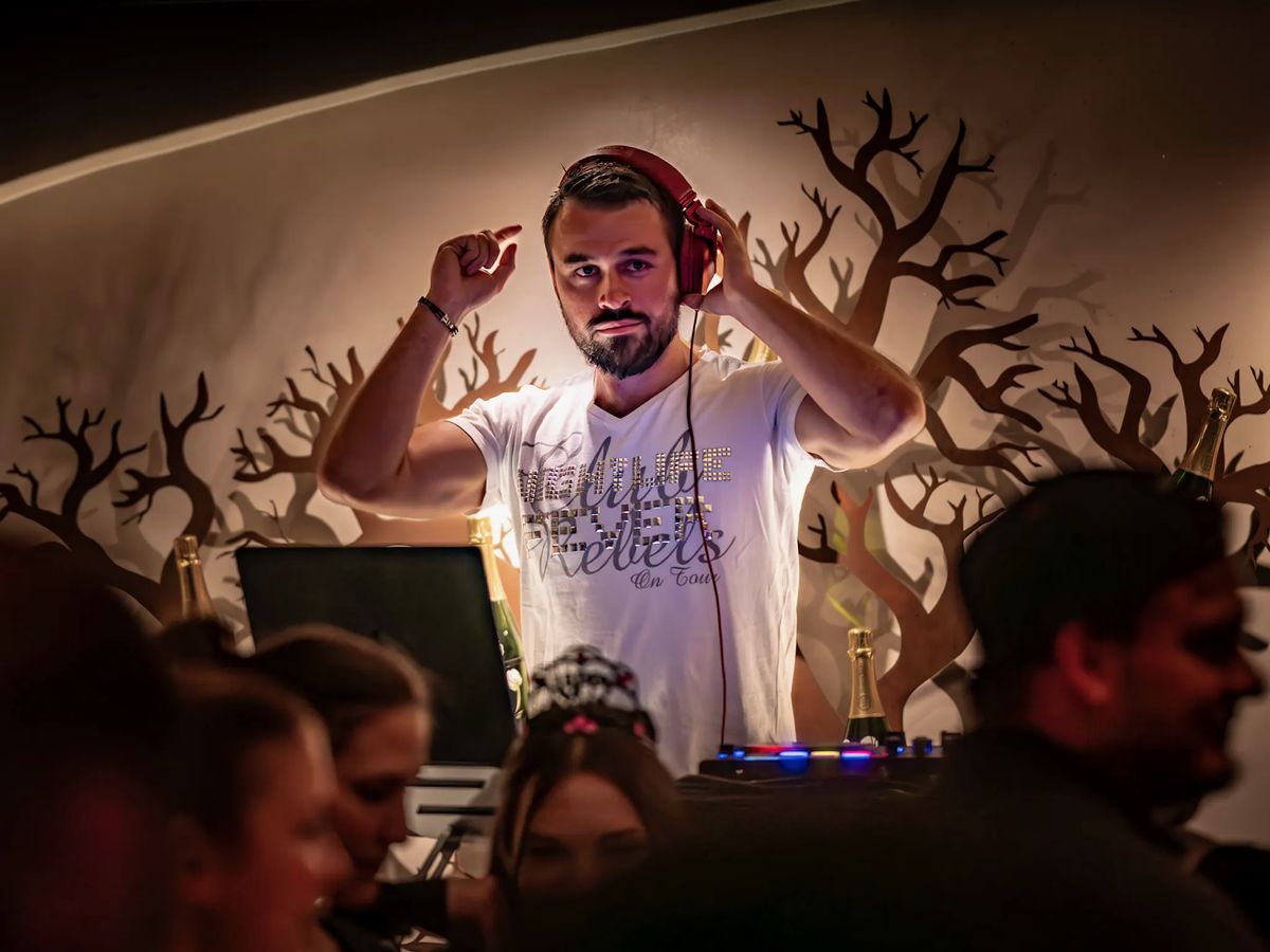 DJ Daniel Wulz setzt am DJ-Pult die Kopfhörer auf, umgeben von Gästen in warmem Licht