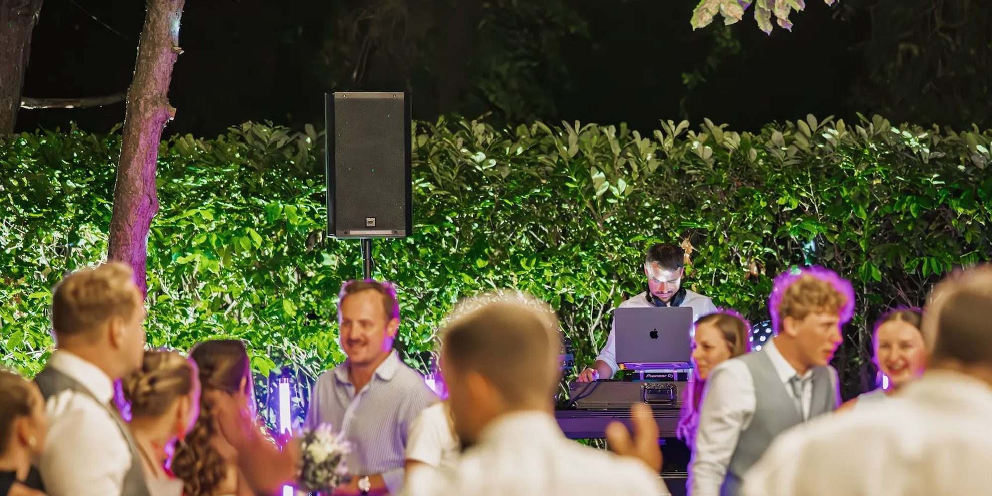 Hochzeitsgäste tanzen Arm in Arm im Kreis vor dem DJ-Setup, das vor einer grünen Hecke aufgebaut ist