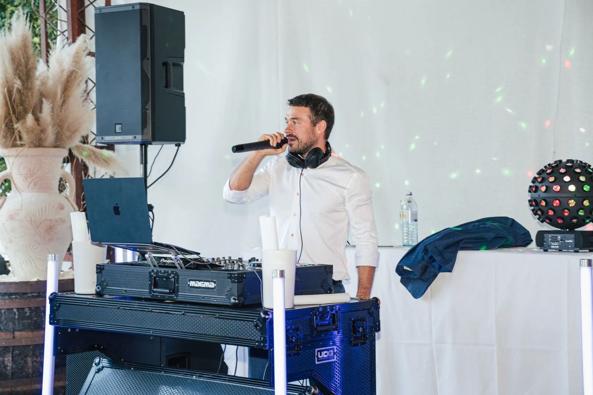 DJ Daniel Wulz im weißen Hemd moderiert mit Mikrofon hinter seinem DJ-Pult