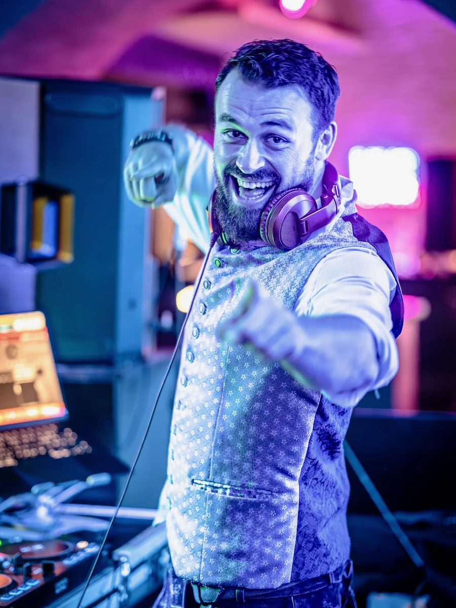 DJ Daniel Wulz in Trachtenweste mit Kopfhörern zeigt lachend vom DJ-Pult in die Kamera